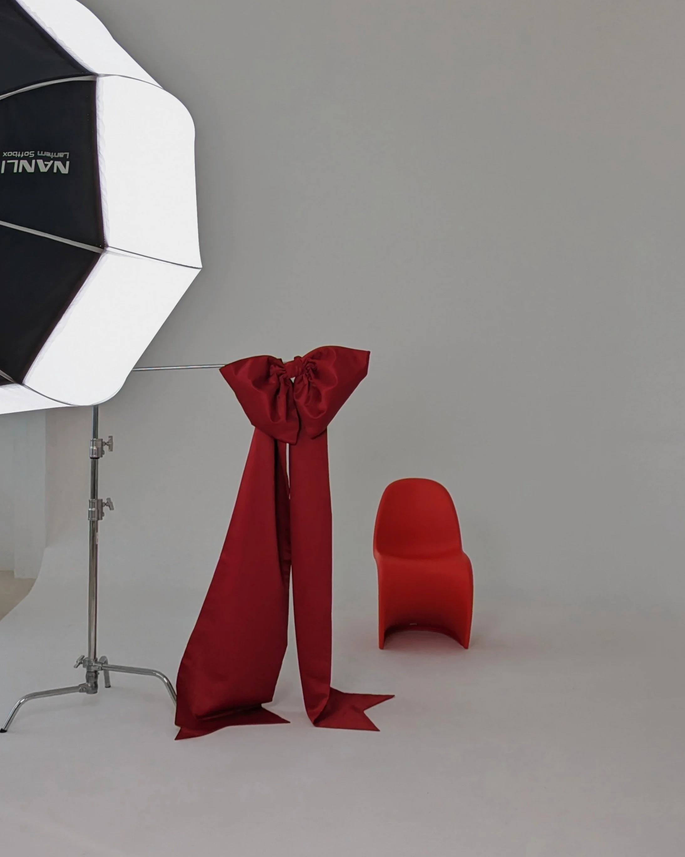 Photography studio setup with an umbrella light, a red cloth tied in a bow, a red chair, all on a white background.