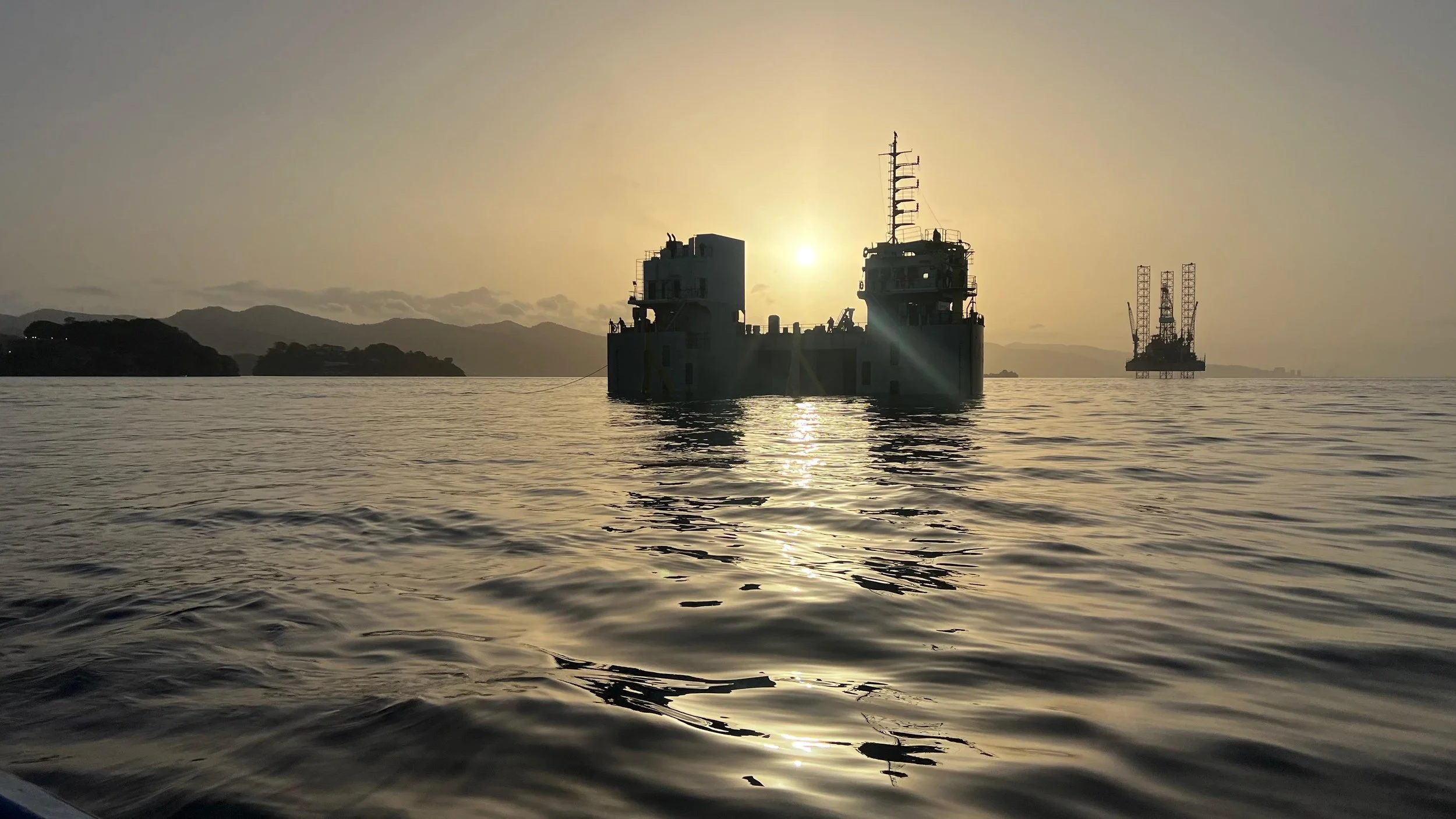 Sun setting behind a large military or research vessel on the water with mountains and a floating platform or drilling rig in the background.