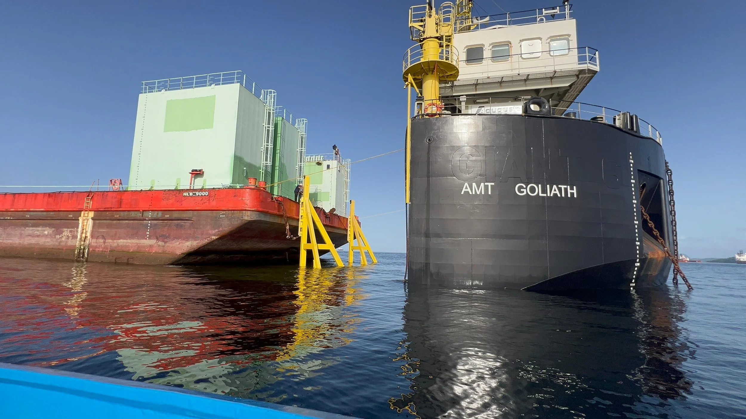 A large black ship named Goliath with yellow and white structures, and a red barge attached to it, floating on calm water under a clear blue sky.