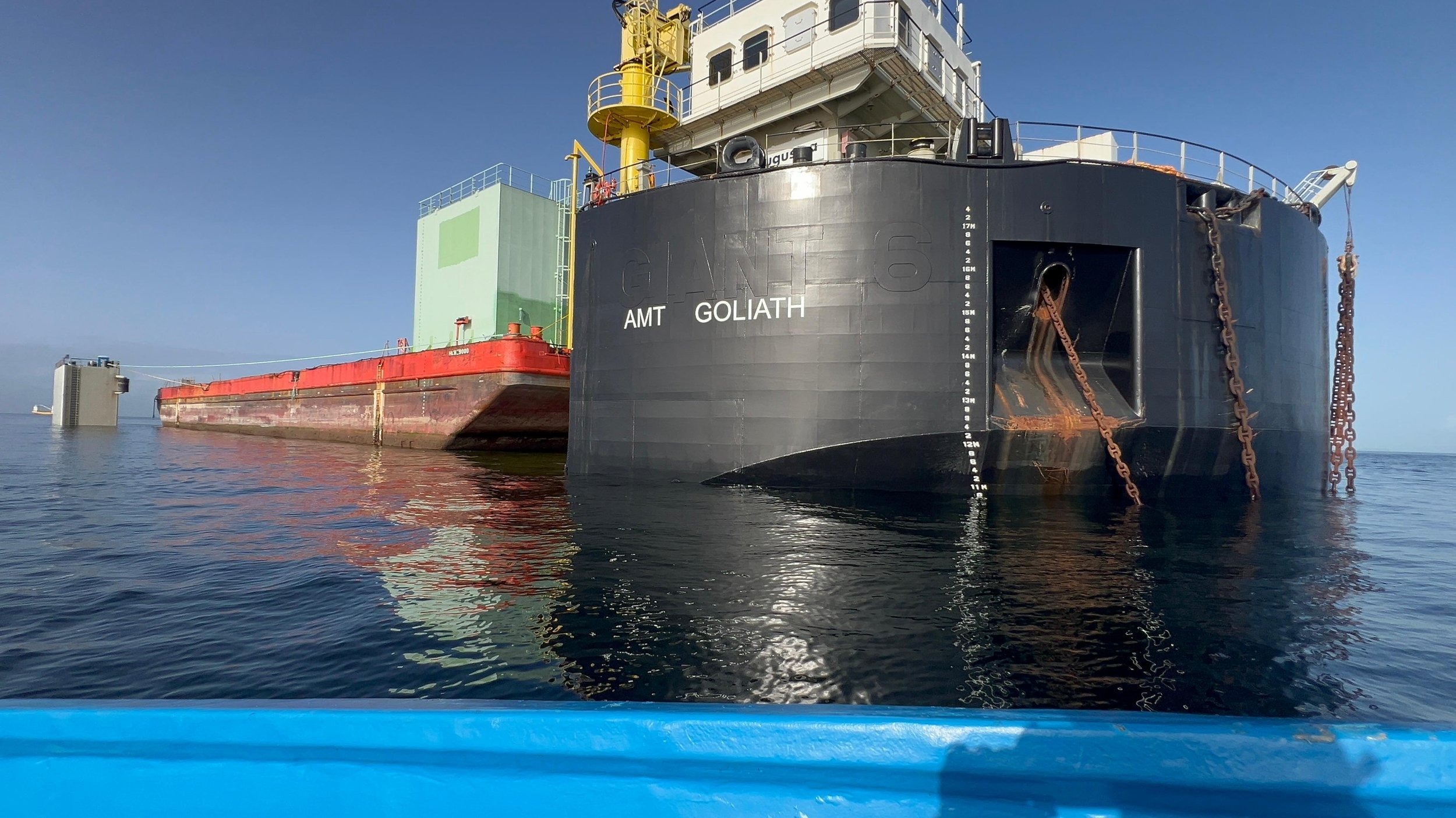 Close-up of a large black cargo ship named GOLIATH with a white superstructure, floating on calm water under clear blue sky. The ship is anchored with chains visible at the bow.