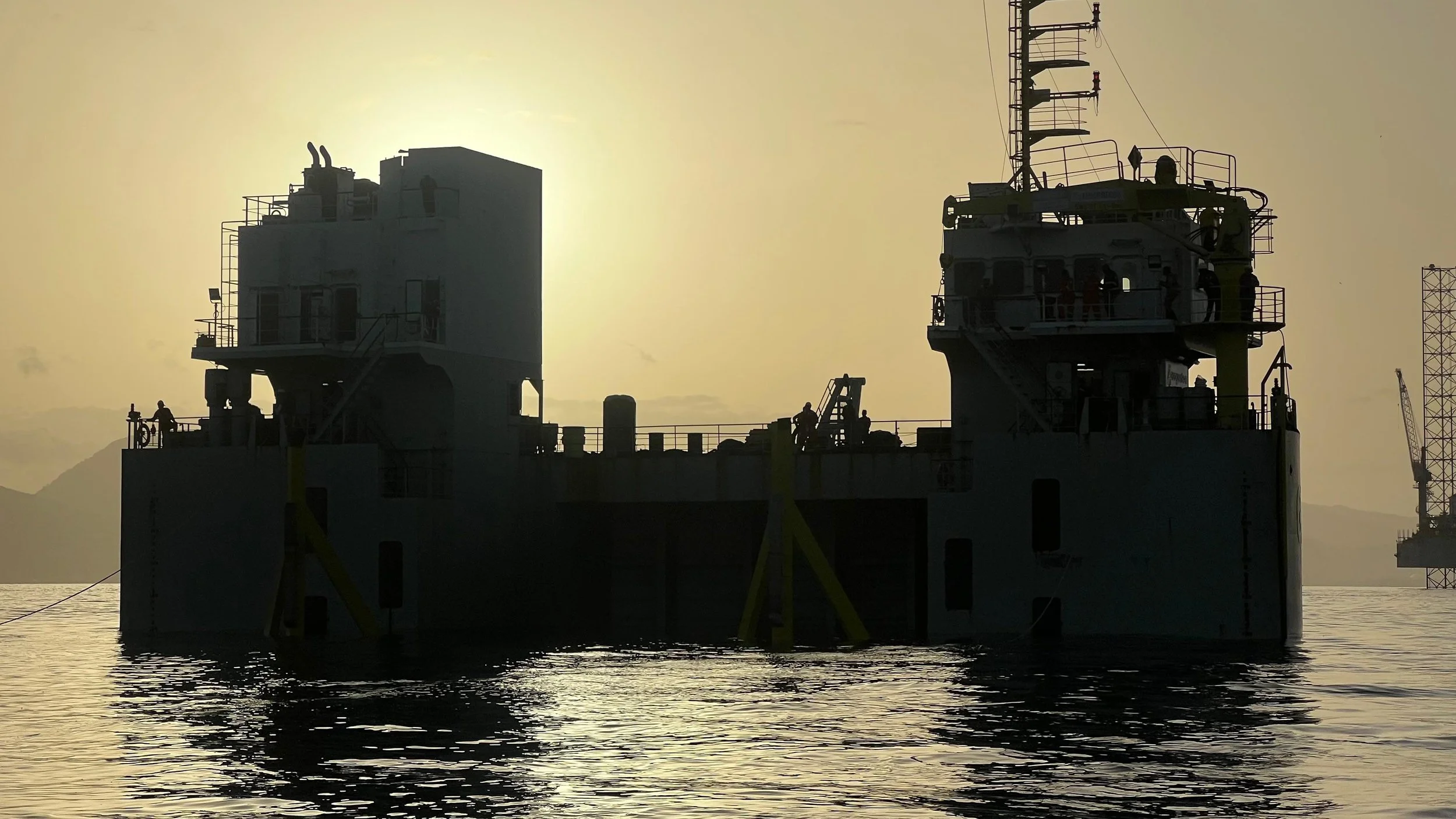 Silhouette of an offshore oil platform or ship at sunset with a calm sea in the foreground.