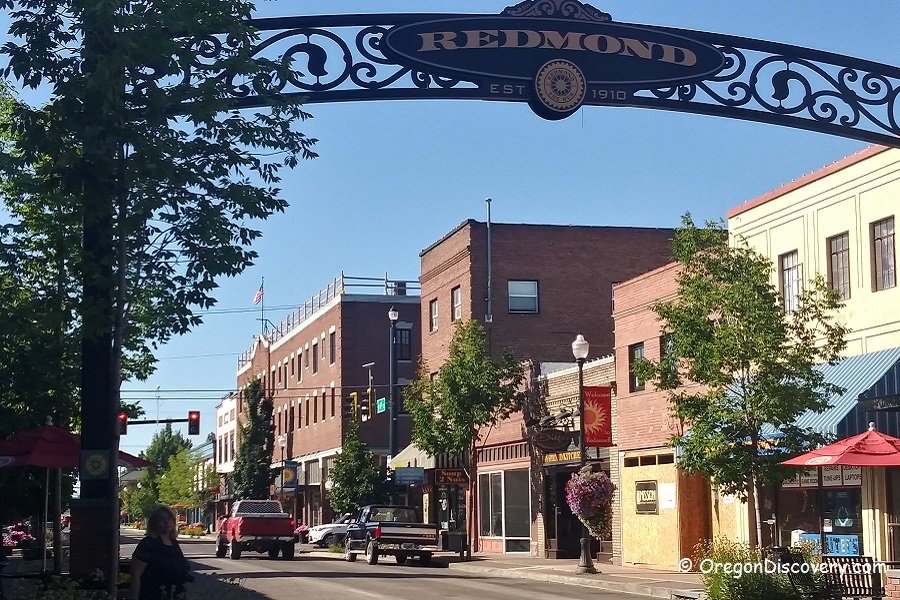 Street view of downtown Redmond, Oregon, with brick buildings, storefronts, trees, outdoor seating with umbrellas, and a decorative sign arch overhead.