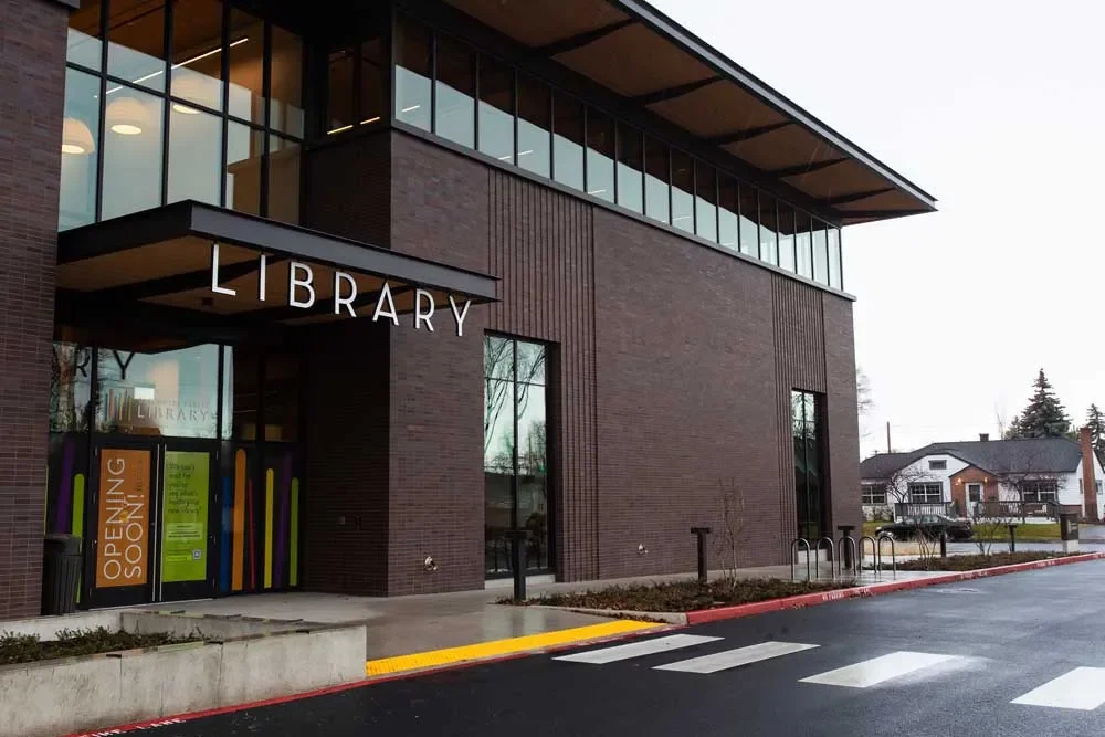 Modern brick library building with glass windows, marked by a sign reading 'LIBRARY' and a poster indicating it is opening soon, located in a suburban area.
