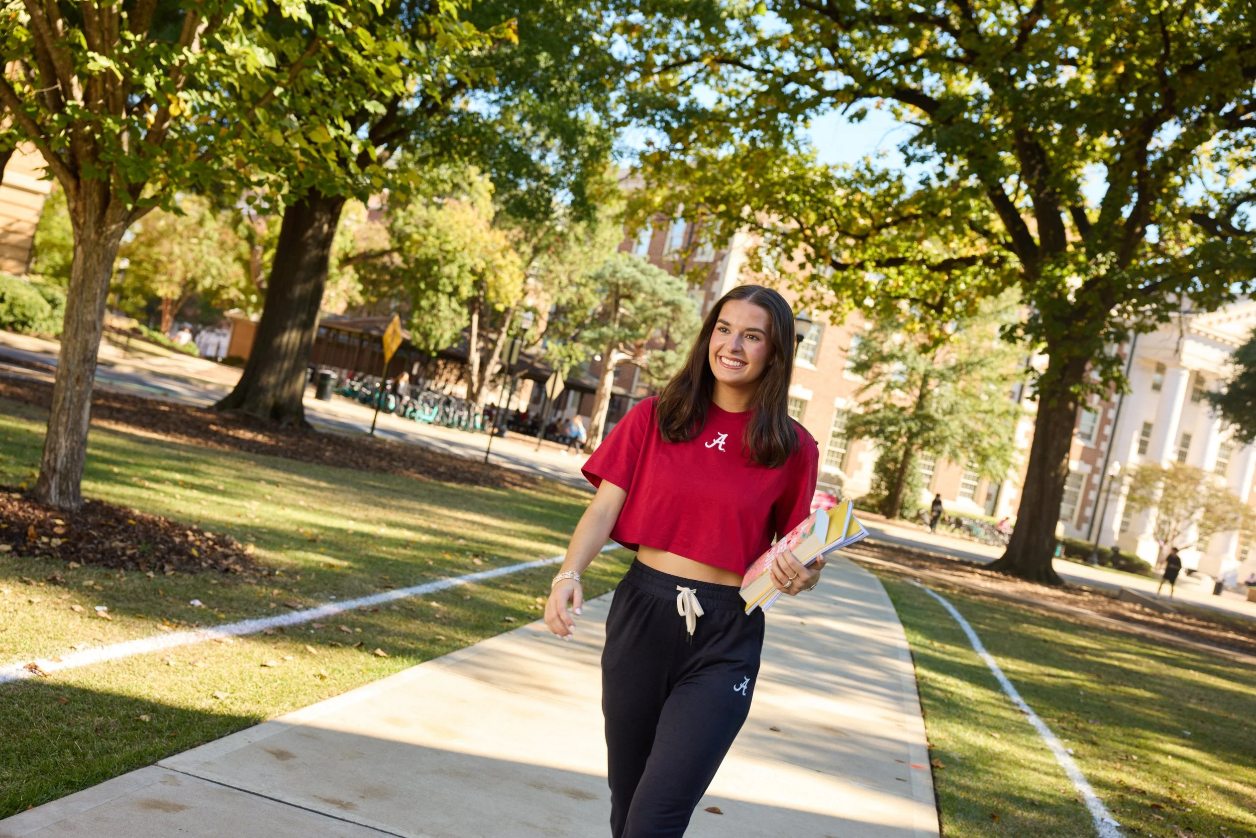 A girl walking on a sidewalk on the University of Alabama campus, holding a stack of books, smiling cheerfully.