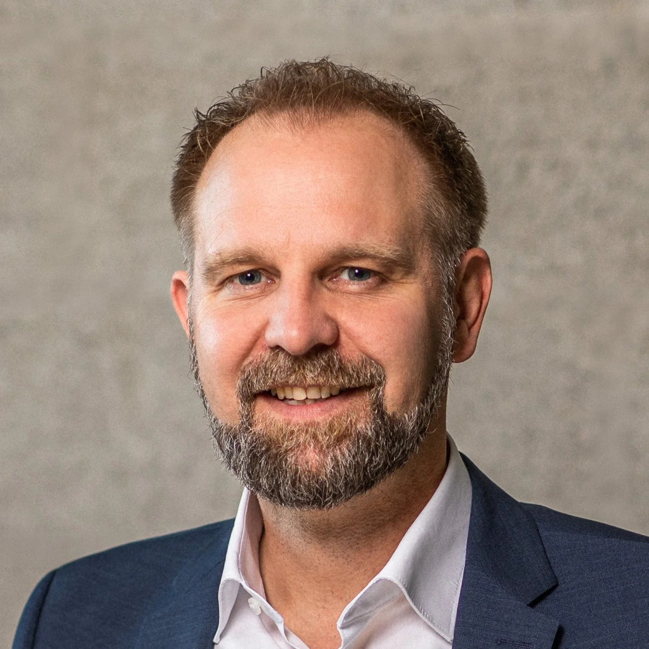 Porträt eines Mannes mit braunem Haar, Bart. Er trägt einen dunkelblauen Anzug und ein weißes Hemd vor einem grauen Hintergrund.