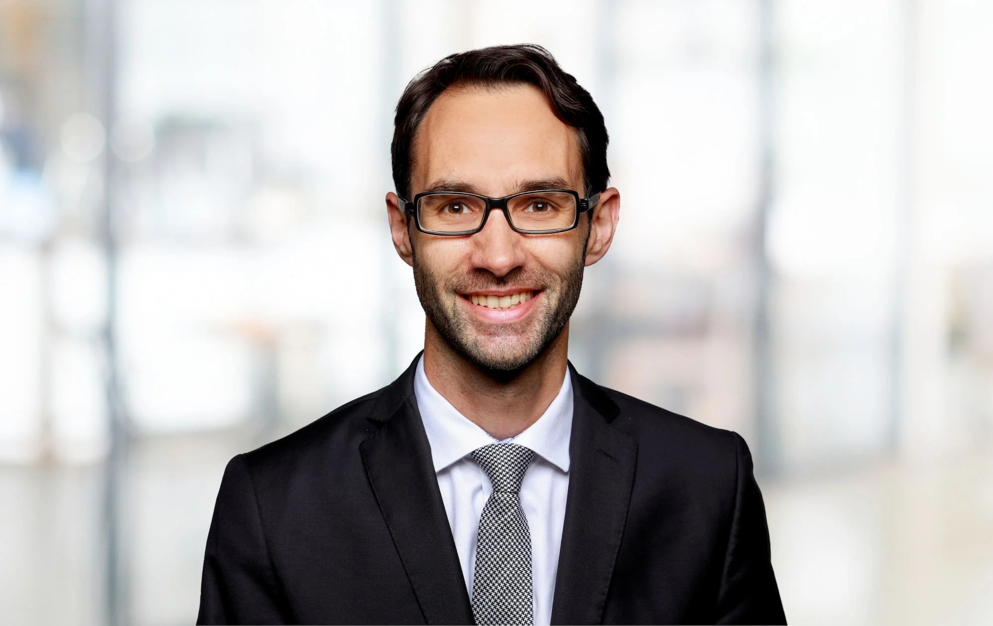 Ein lachender Mann in einem schwarzen Anzug, weißem Hemd und Silberkrawatte, trägt Brille, vor einem unscharfen hellen Hintergrund.