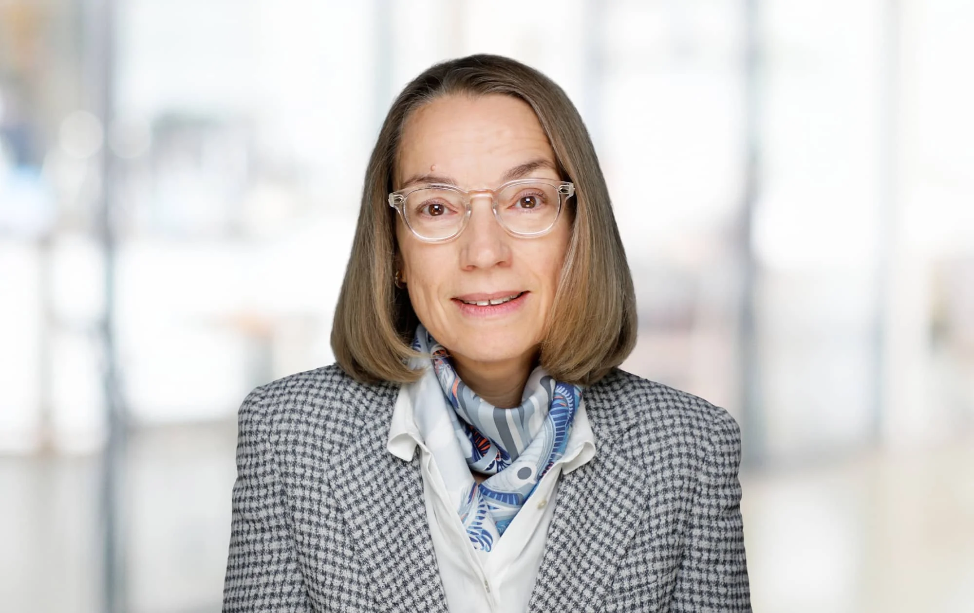 Eine Frau mit Brille, mittellangem braunen Haar und einem Blazer, die vor einem Bürohintergrund steht.