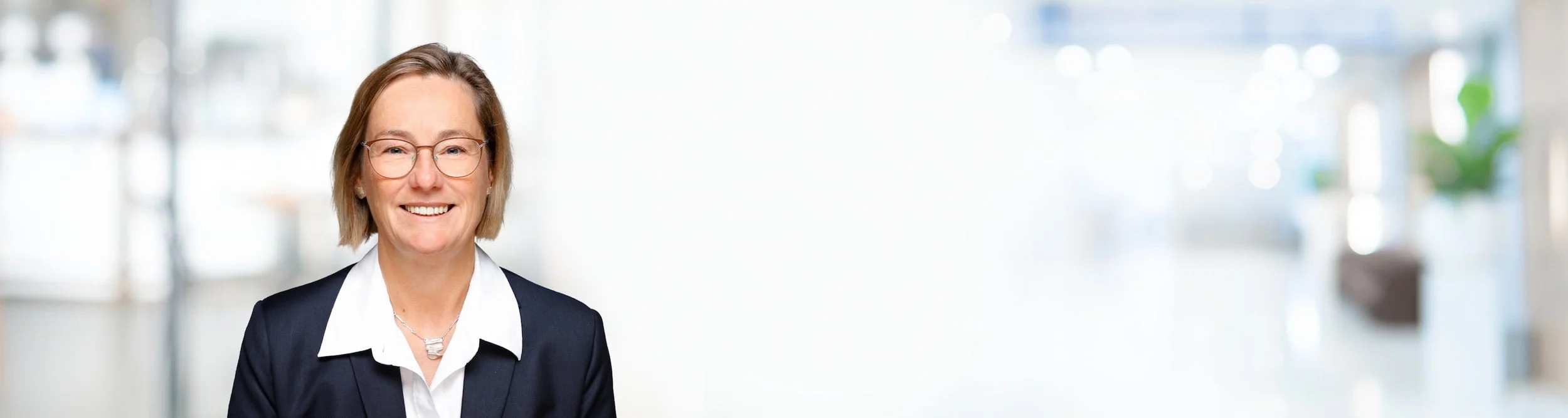 Eine smiling Frau mit Brille in einem dunklen Blazer und weißen Hemd vor einem hellen Hintergrund.