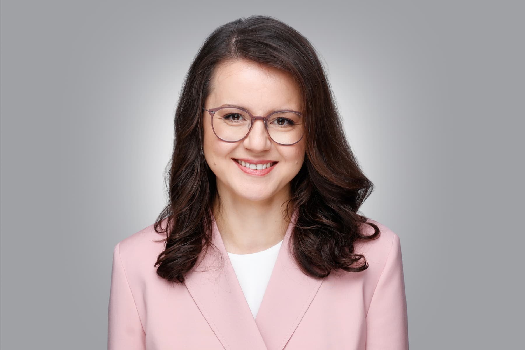 Porträt von Nina Ayvazova-Mosch mit dunkelbraunem Haar, Brille, hellrosa Blazer und weißer Bluse vor hellgrauem Hintergrund.