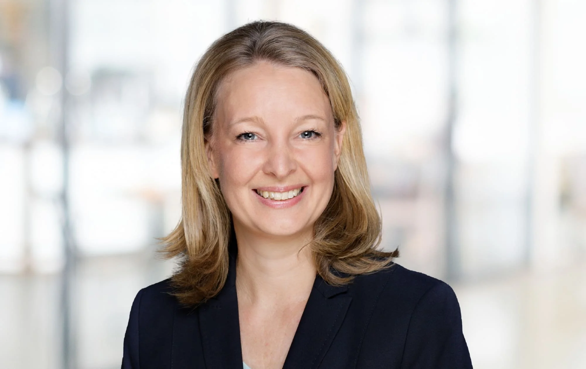Professionelles Porträt einer lächelnden Frau mit mittellangen, gewellten, hellbraunen Haaren vor einem hellen, unscharfen Hintergrund.