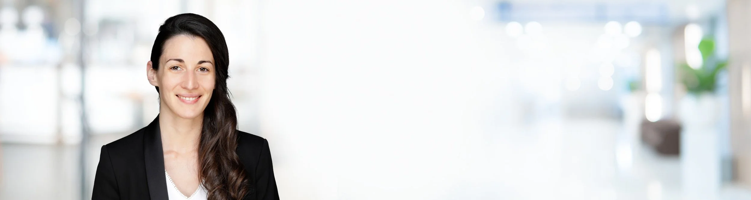 Eine lachende Frau mit dunklem, schulterlangem Haar in einem schwarzen Blazer vor einem hellen, unscharfen Hintergrund, vermutlich in einem Büro.