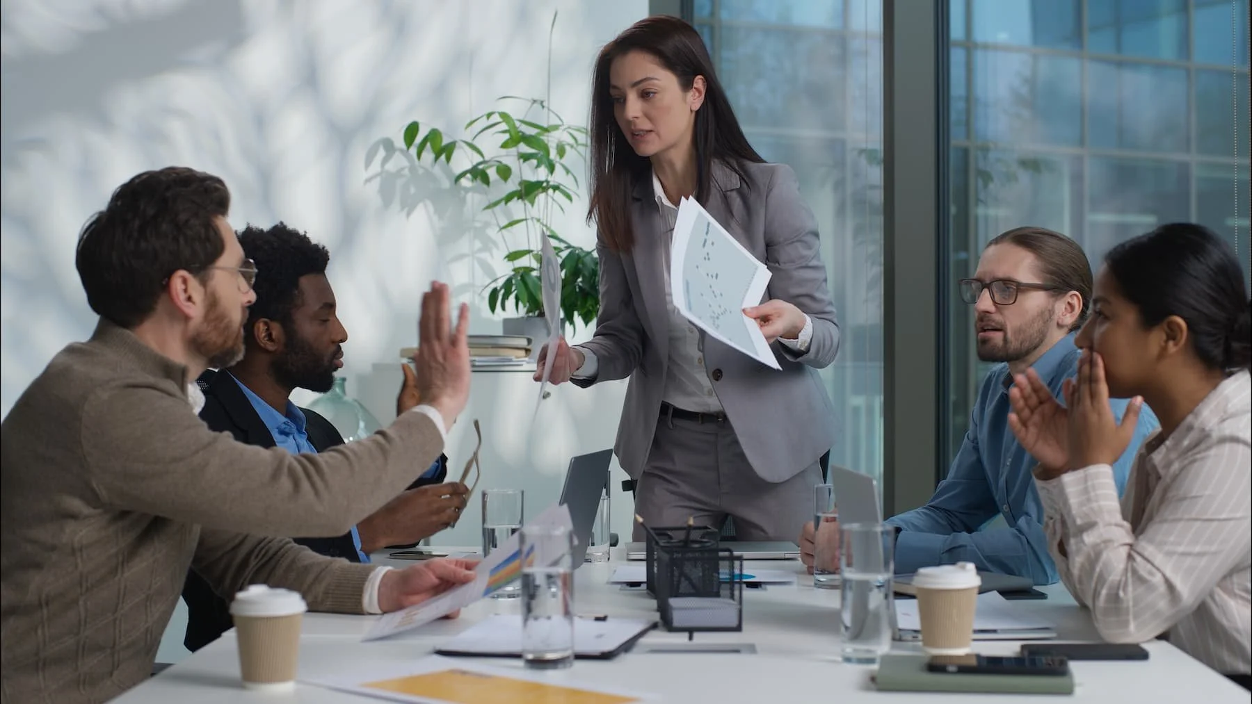 Geschäftsbesprechung in einem modernen Büro: Eine Frau im Business-Outfit verteilt Unterlagen an mehrere Mitarbeitende während einer Diskussion über Sicherheits- oder Compliance-Themen.