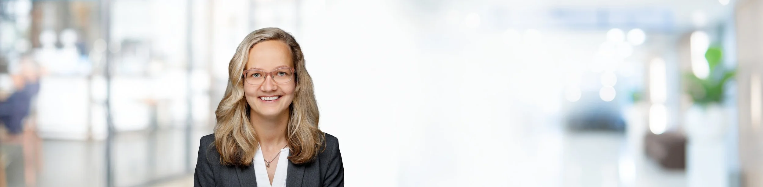 Porträt einer lächelnden jungen Frau mit Brille vor einem unscharfen Bürohintergrund.