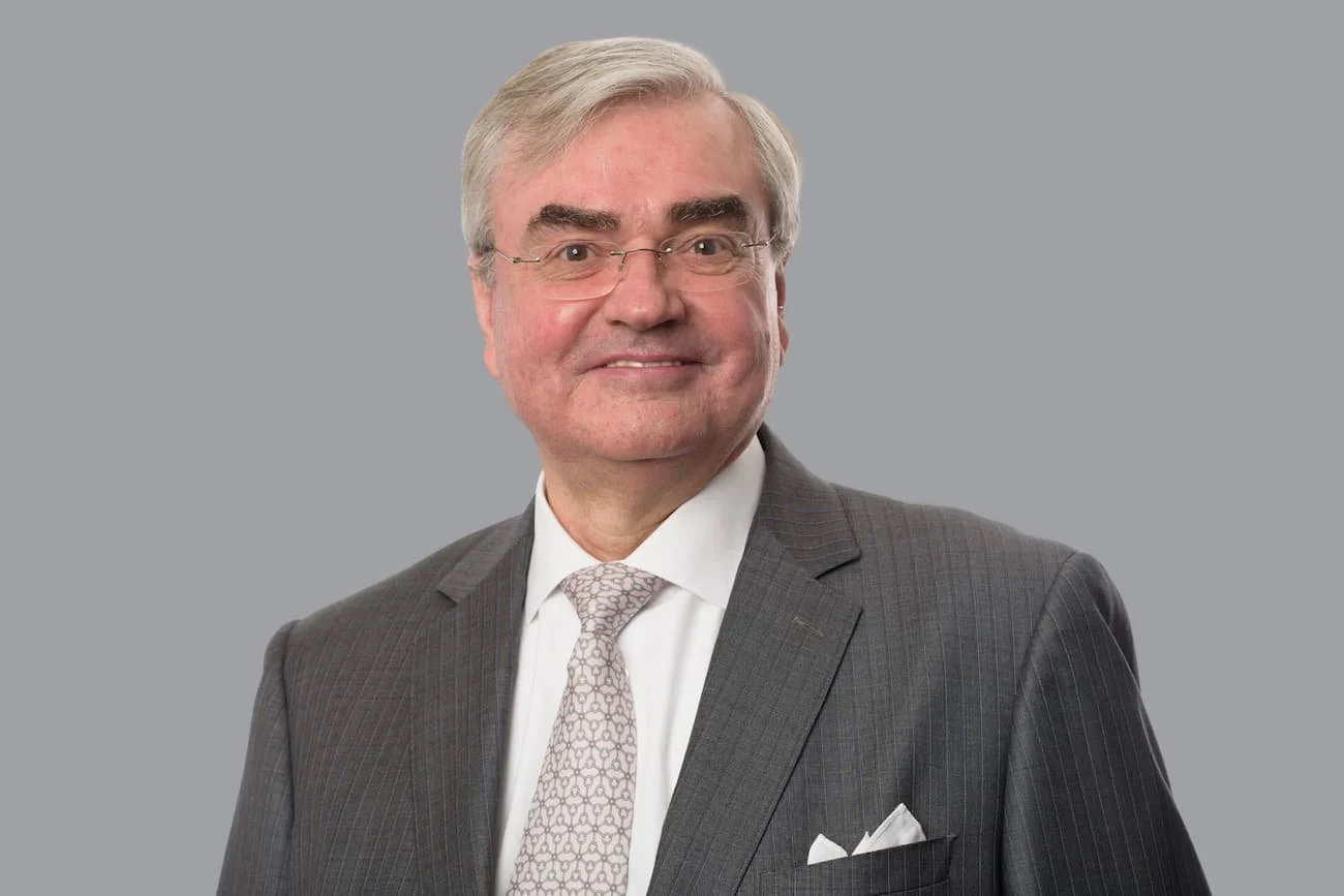 Ein lächelnder Mann in einem grauen Anzug und einer weißen Krawatte vor einem grauen Hintergrund.