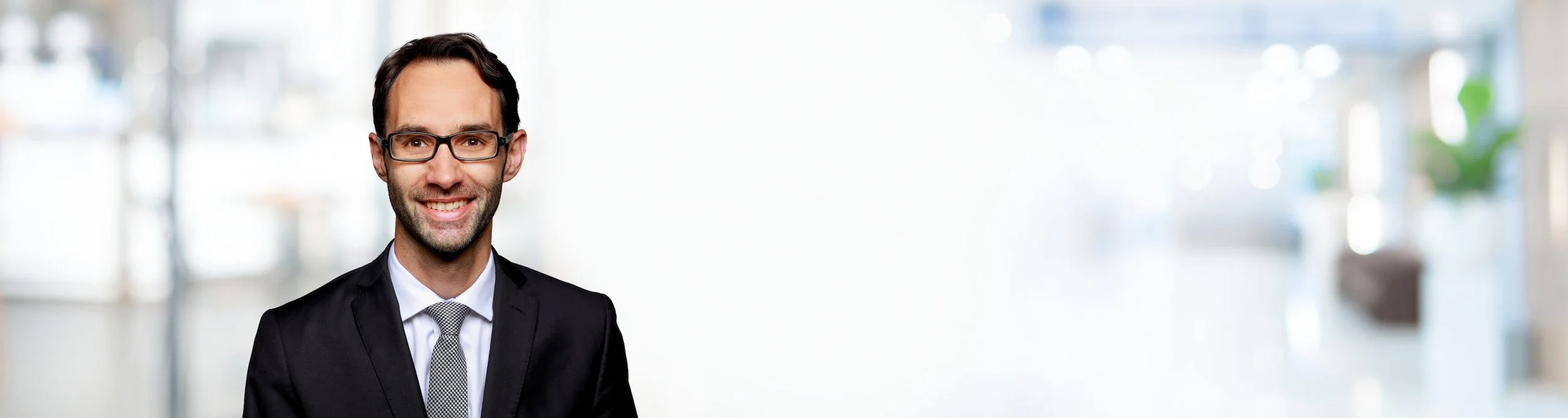 Ein lächelnder Mann in einem schwarzen Anzug, weißem Hemd und grauem Krawatte, trägt Brille, vor einem hellen, unscharfen Hintergrund.
