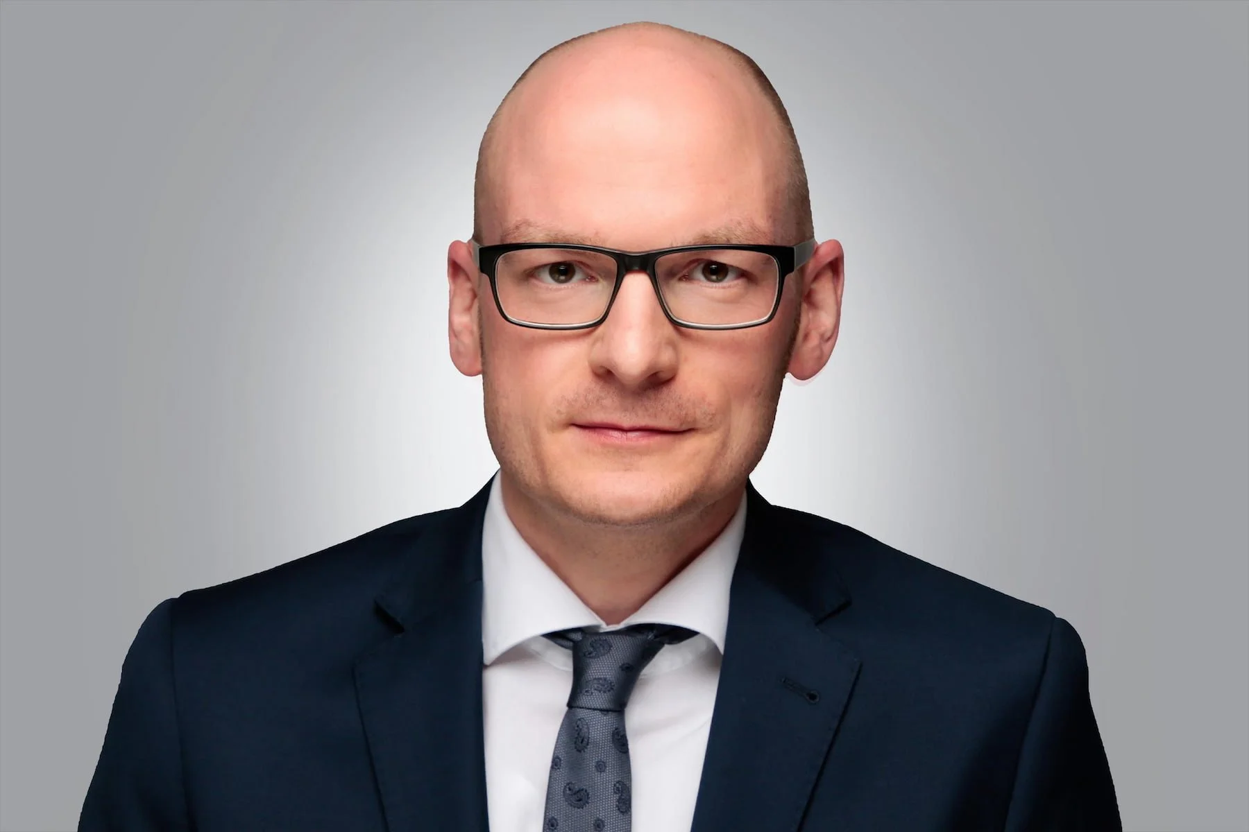 Porträt eines Mannes in formeller Kleidung mit Brille vor grauem Hintergrund.