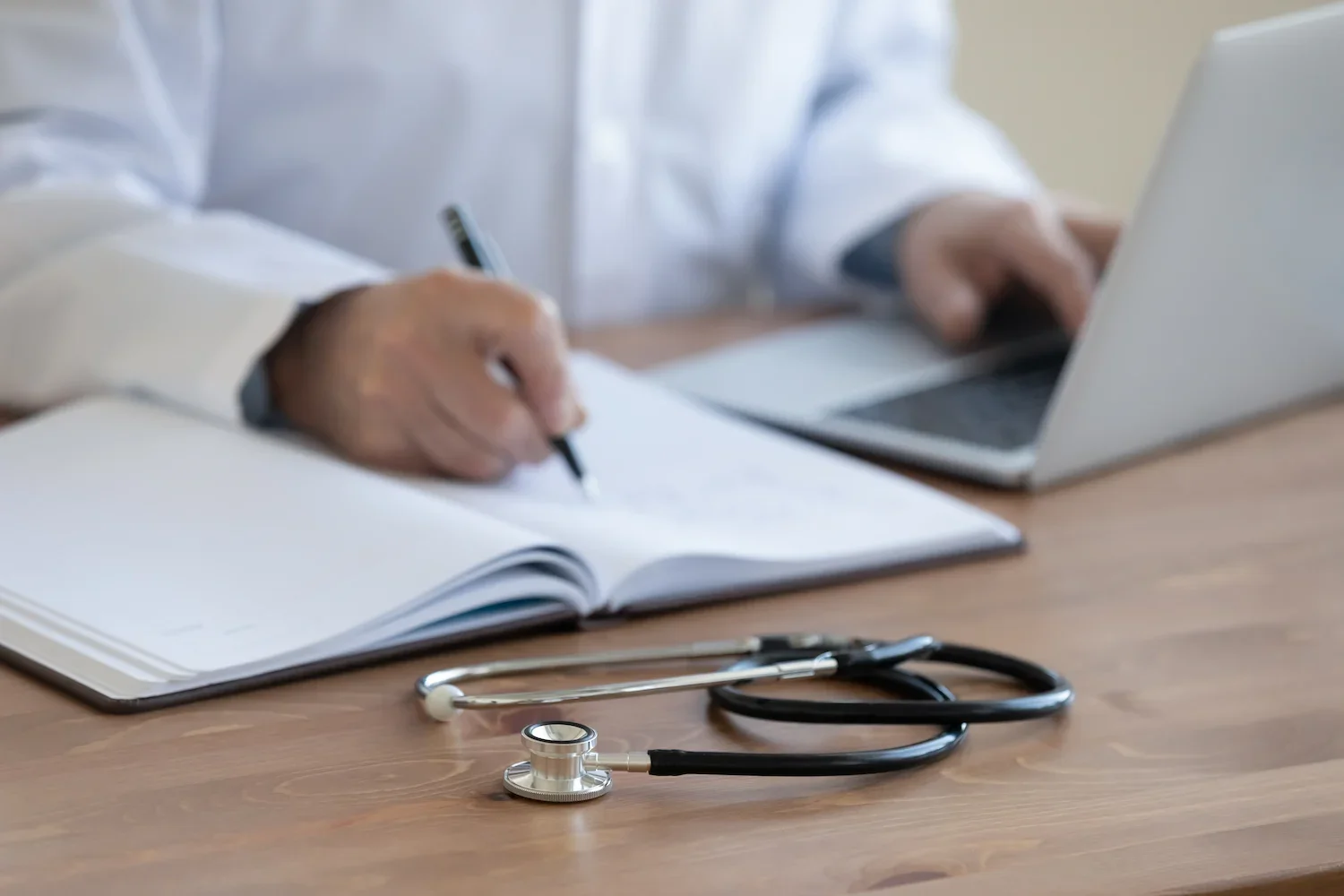 Ärztinnen- oder Arzthände mit Stethoskop, Notebook, Notizbuch und Schreibgerät auf einem Holztisch.