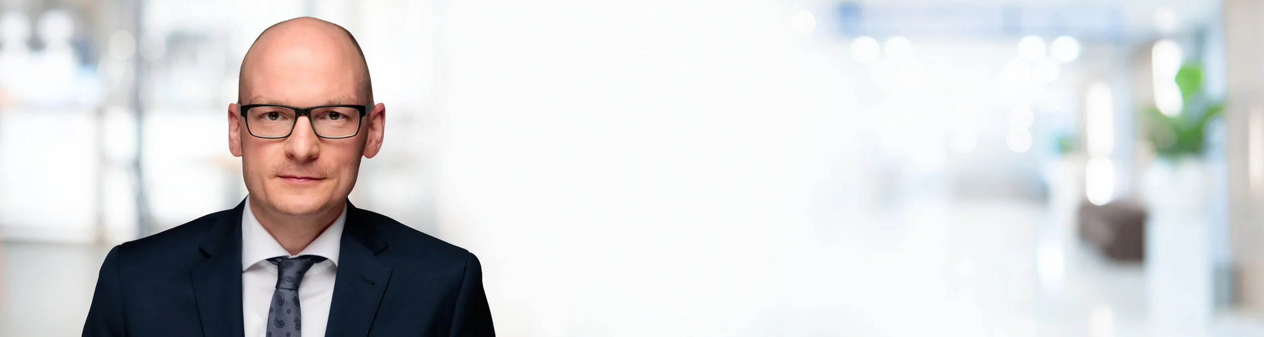 Ein Mann in einem dunklen Anzug mit Krawatte und Brille vor einem hellen, unscharfen Hintergrund.