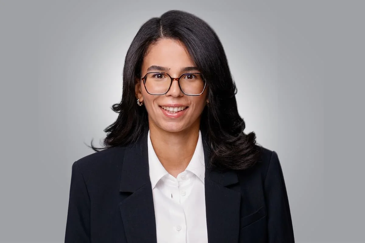 Porträt einer lächelnden Frau mit schwarzen Haaren, Brille, dunklem Blazer und weißem Hemd vor grauem Hintergrund.