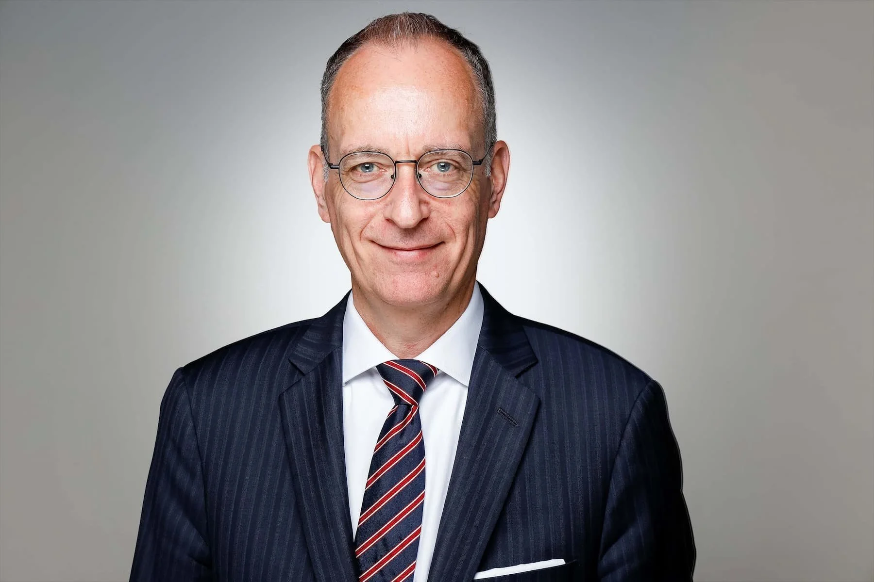 Ein Mann in einem dunklen Anzug mit einer weißen Hemd und gestreiftem Krawatte, trägt Brille und lächelt. Hintergrund ist grau und schlicht.