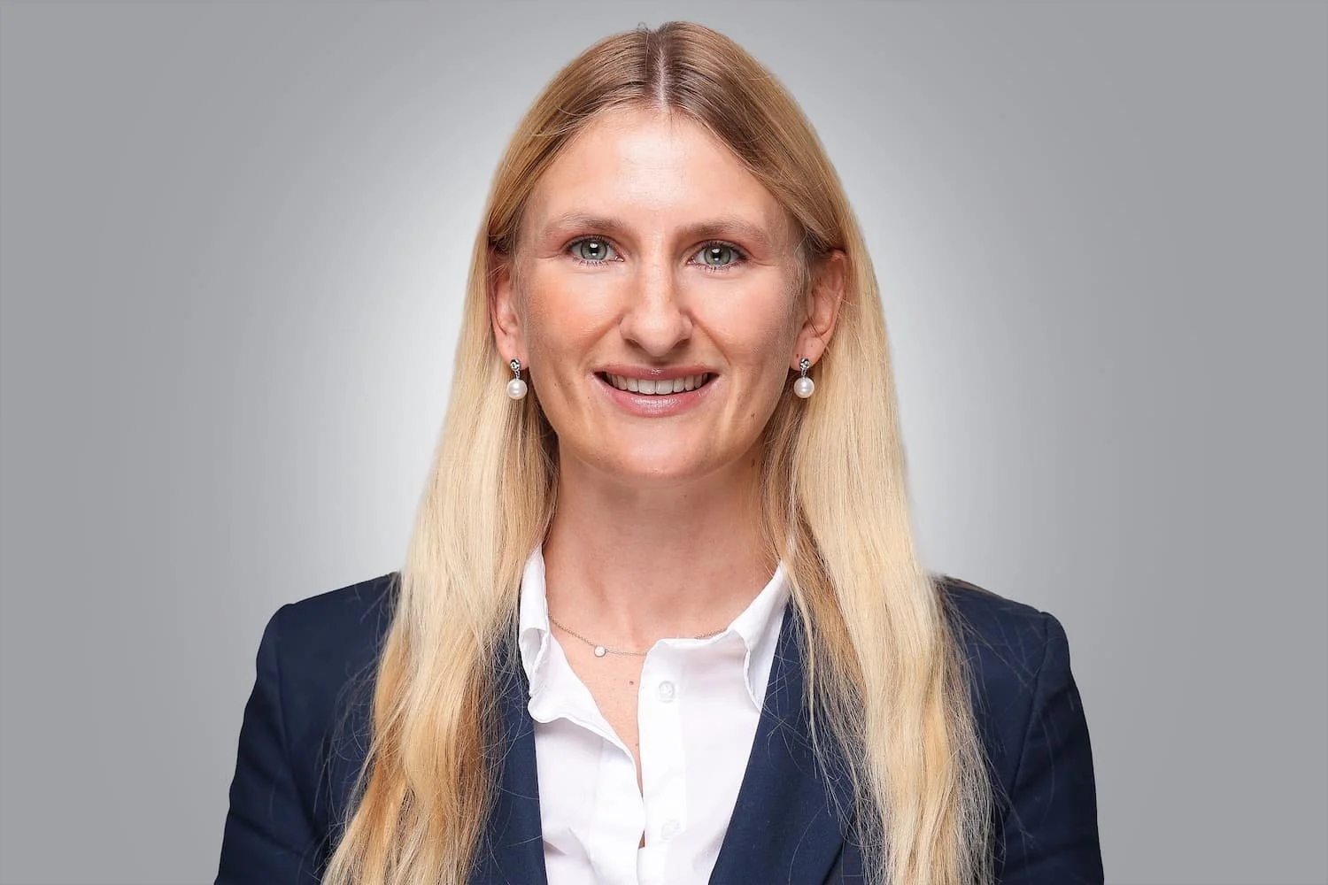 Porträt einer Frau mit blonden Haaren, blauen Augen, in Businesskleidung und Perlenohrringen vor neutralem Hintergrund.