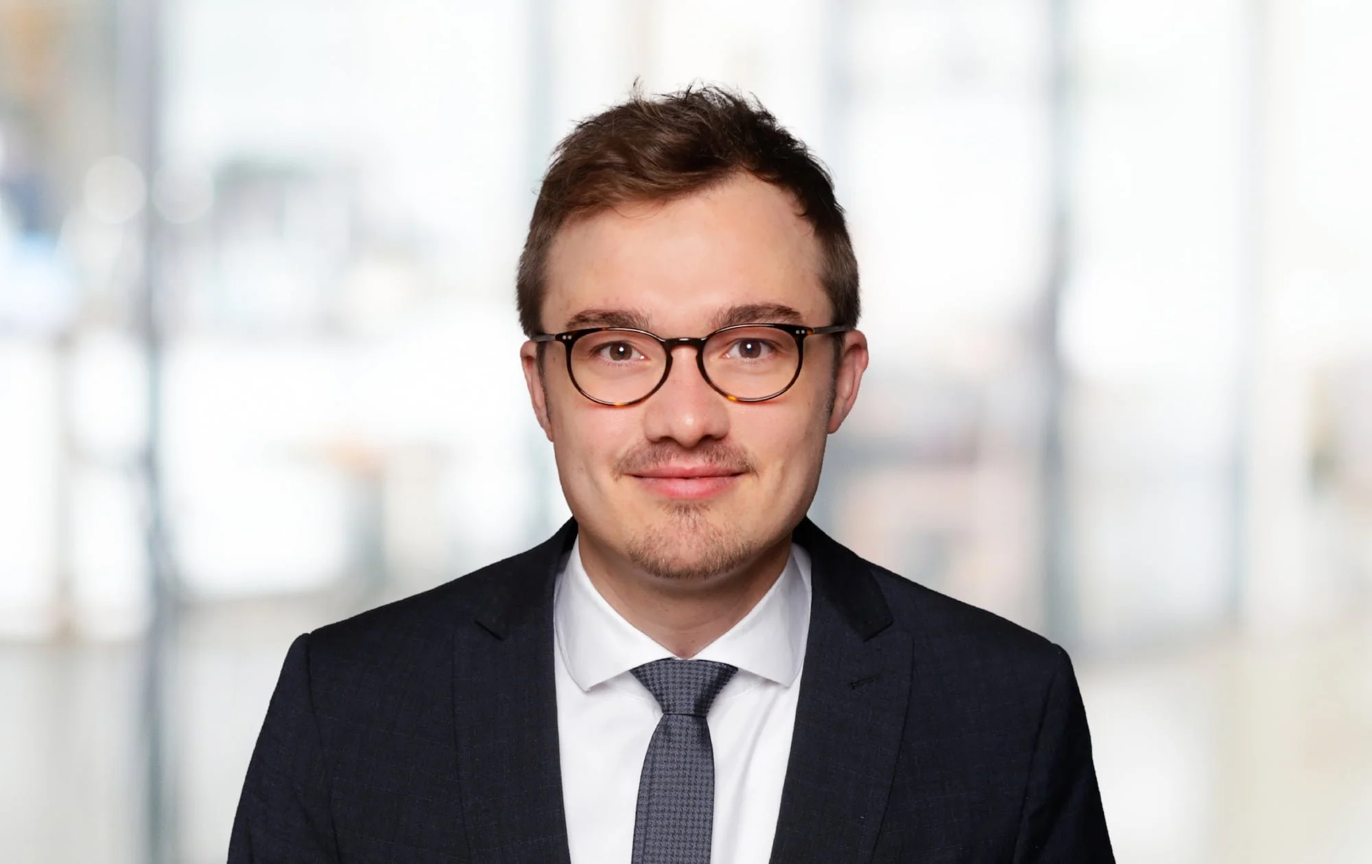 Porträt eines jungen Mannes in formeller Kleidung, trägt Brille, vor verschwommenem Büro-Hintergrund.