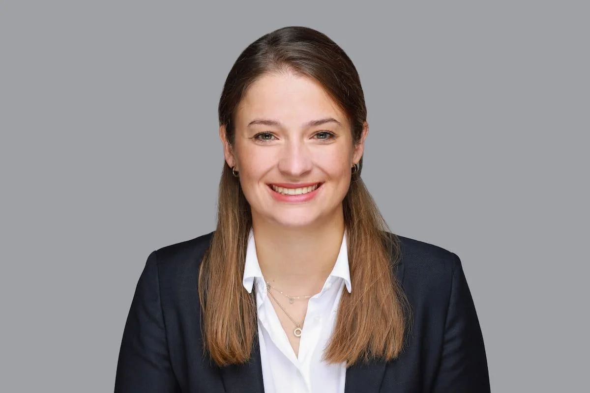 Porträt einer Anwältin im dunklen Blazer und weißer Bluse, freundlich lächelnd vor neutralgrauem Hintergrund.