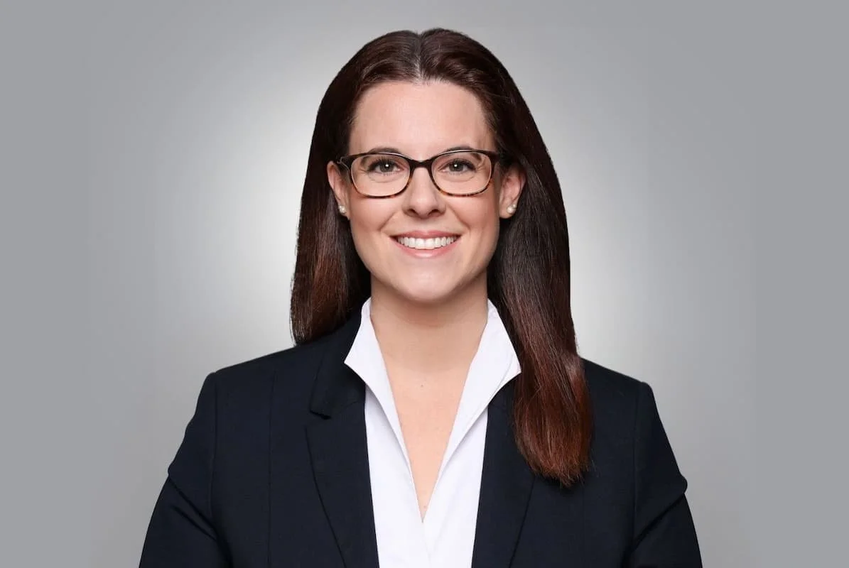 Porträt einer Frau im dunklen Blazer und weißer Bluse mit Brille, freundlich lächelnd vor hellem, neutralem Hintergrund.