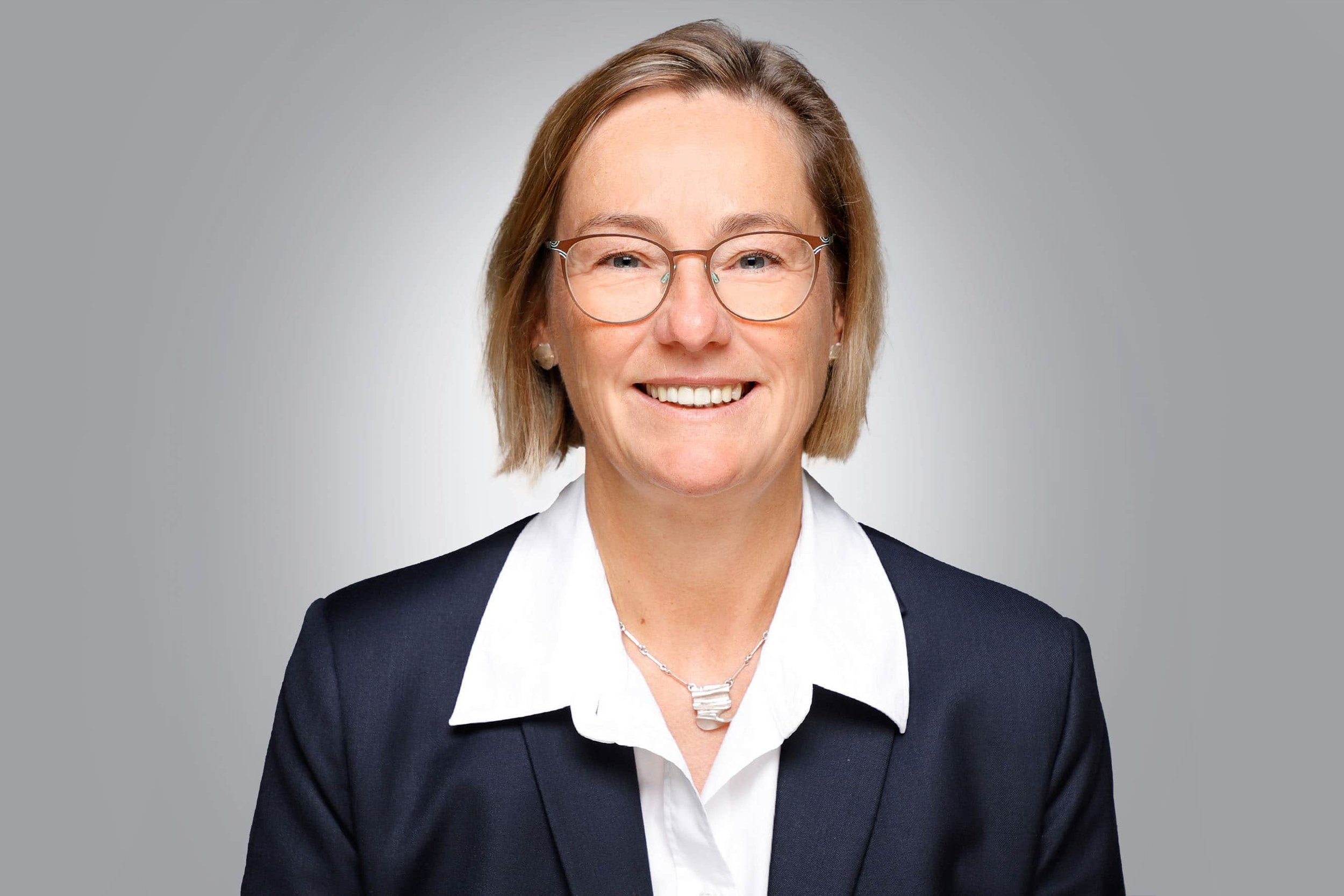 Porträt einer Frau mit Brille, kurzes Haar, in formeller Kleidung, vor grauem Hintergrund.