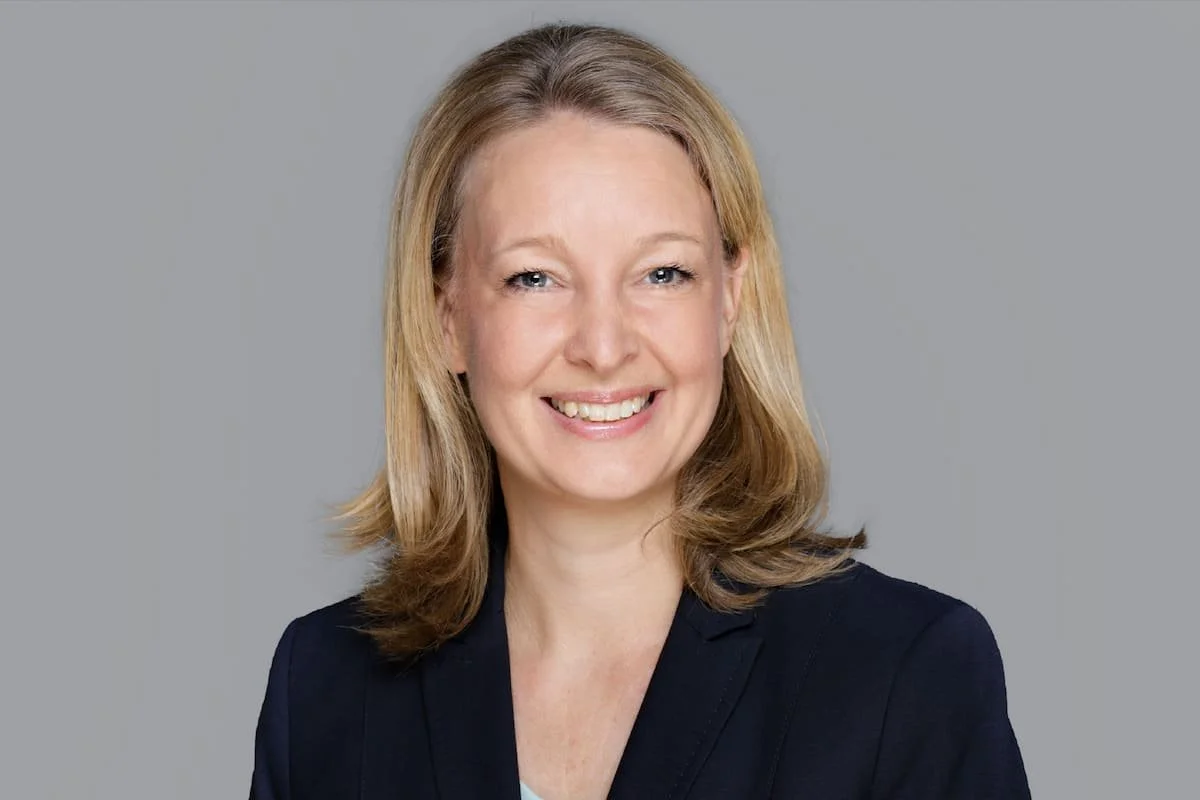 Porträt einer lächelnden Frau mit blonden Haaren, die schwarze Jacke trägt, vor einem grauen Hintergrund.