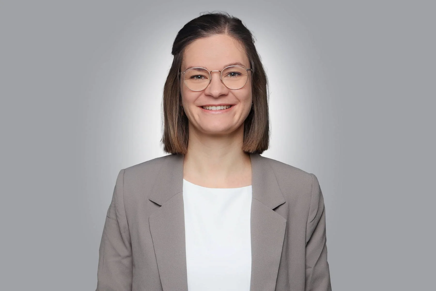 Professionelles Portrait einer jungen Frau mit Brille, braune Haare, grauem Blazer und weißem Hemd vor grauem Hintergrund.