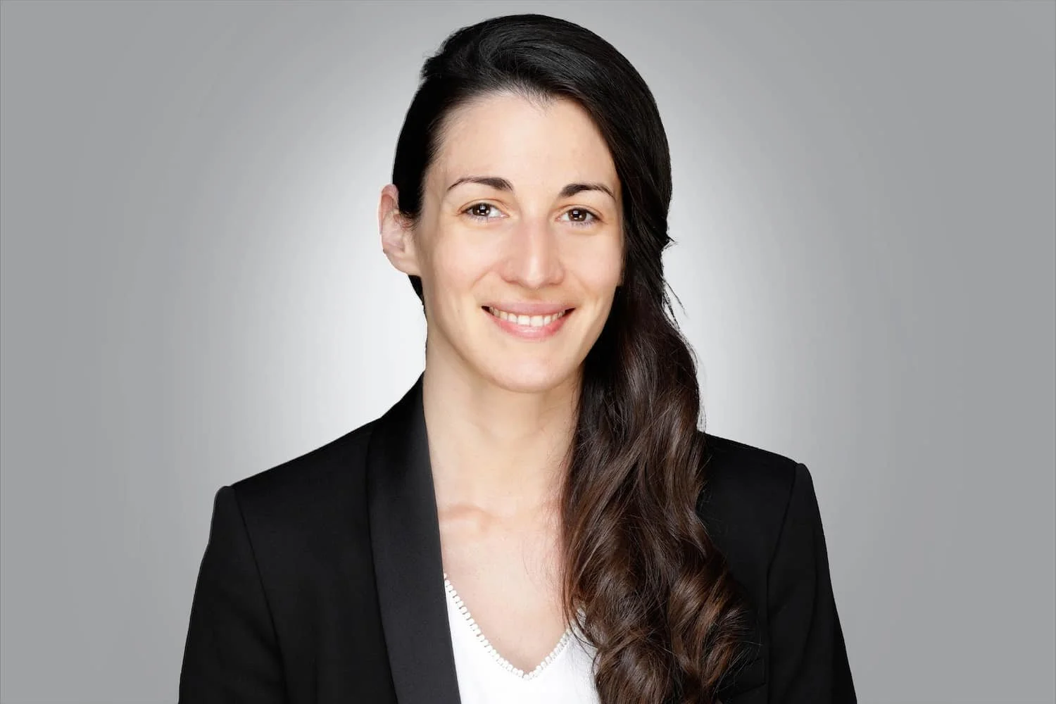 Portrait einer Frau mit langem, braunem, gewelltem Haar, schwarzer Jacke, weißem Top, auf grauem Hintergrund, lächelnd.