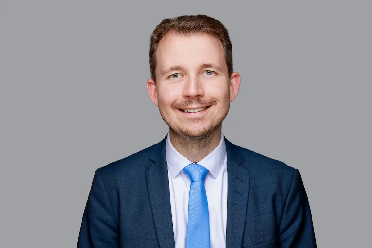 Ein junger Mann trägt einen dunklen Anzug, weißes Hemd und blaue Krawatte und lächelt vor einem grauen Hintergrund.