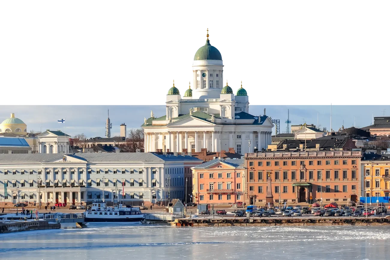 Stadtansicht mit Wasser im Vordergrund, historischen Gebäuden und einer Kirche mit grünen Kuppeln im Hintergrund, vermutlich in Helsinki, Finnland.