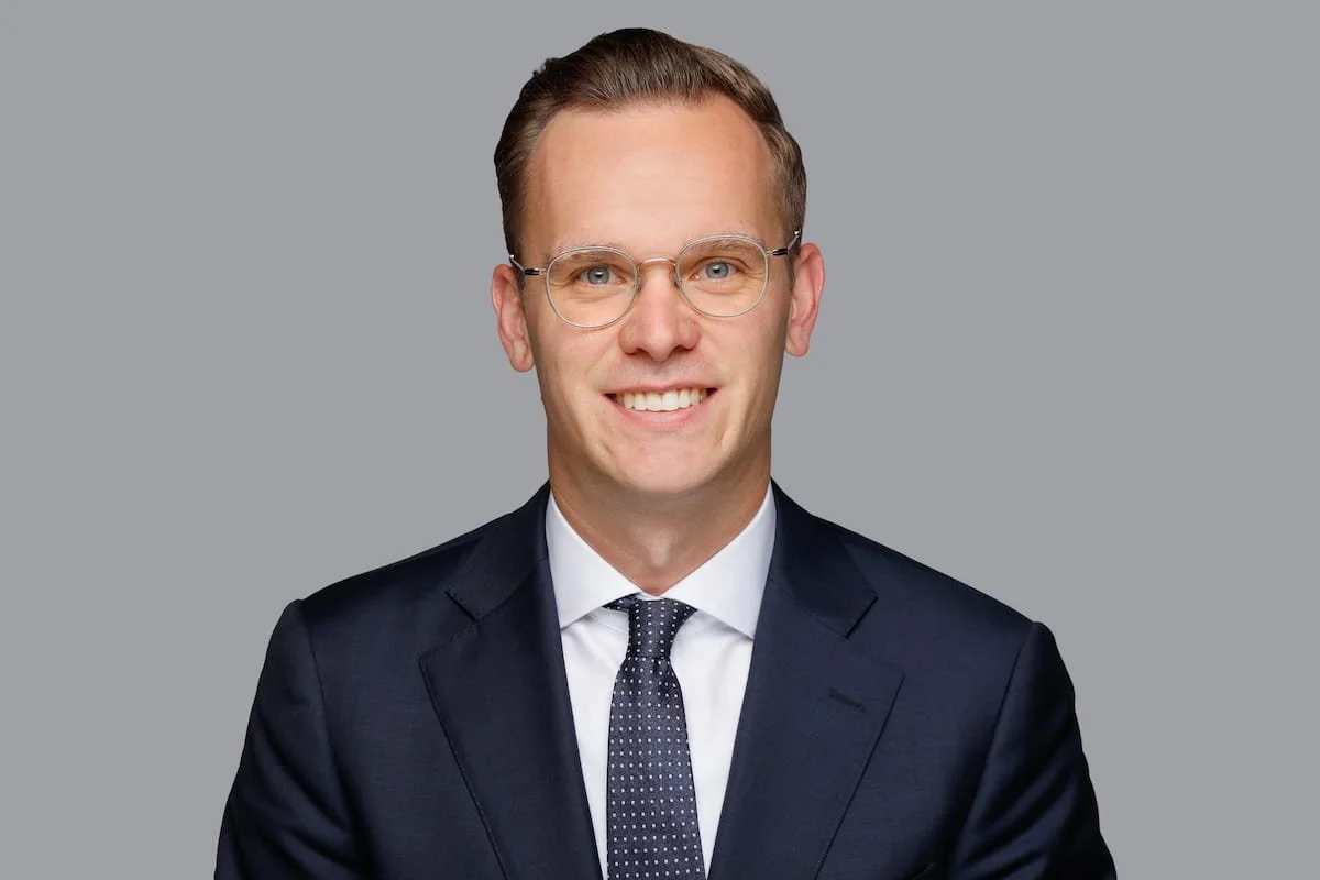 Ein Mann trägt eine dunkle Anzugjacke, ein weißes Hemd und eine dunkelblaue Krawatte mit weißen Punkten. Er trägt runde Brillen und hat kurze, braune Haare. Hintergrund ist grau.