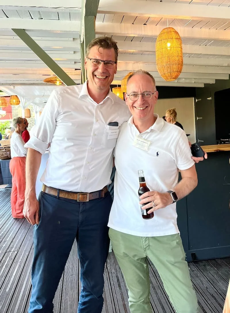 Zwei lachende Männer in einem hellen, modern eingerichteten Raum bei einem Treffen oder einer Feier, einer hält ein Bier, beide tragen Brillen und weiße Hemden.