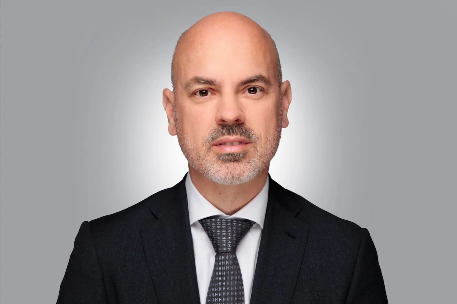 Porträt eines Mannes in formeller Kleidung mit Blond- und Grauhaar, er trägt einen schwarzen Anzug, weißes Hemd und eine schwarze Krawatte, gegen grauen Hintergrund.