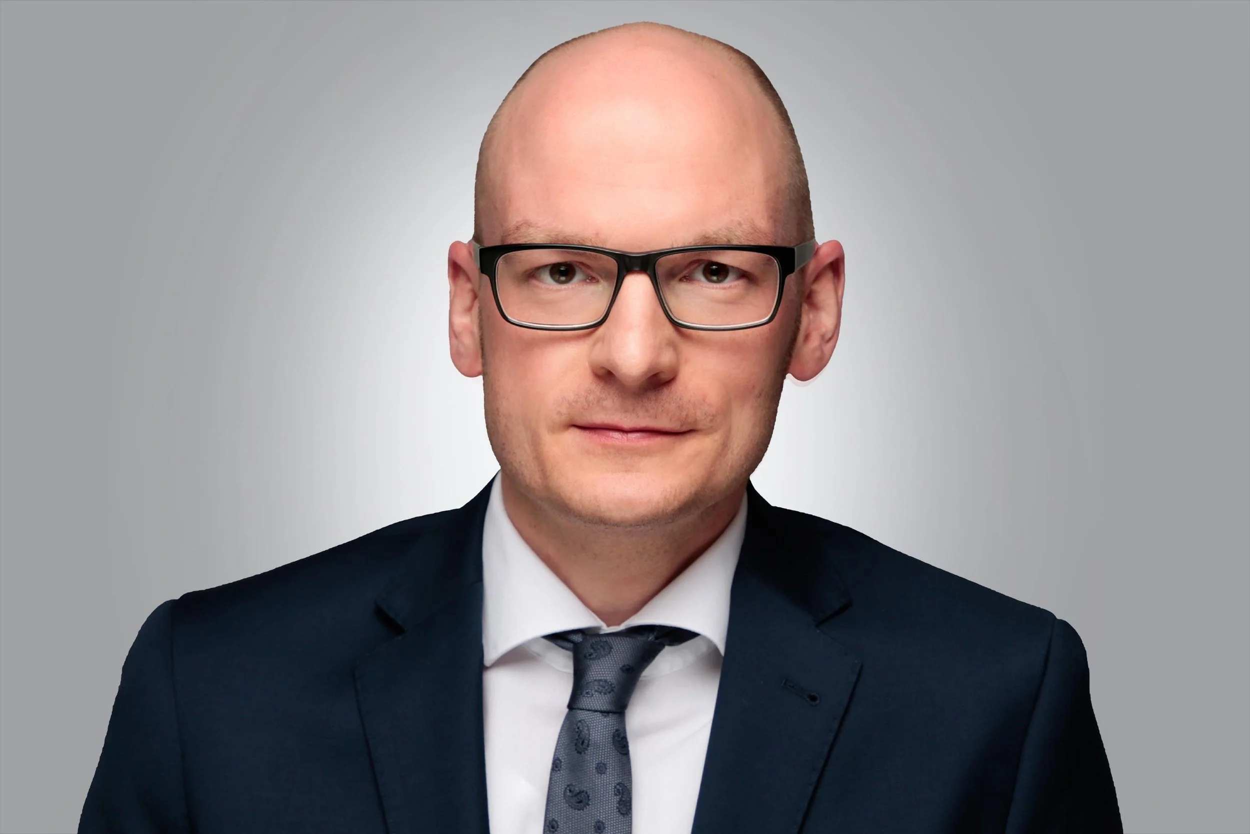 Porträt eines Mannes in formeller Kleidung mit Brille vor grauem Hintergrund.