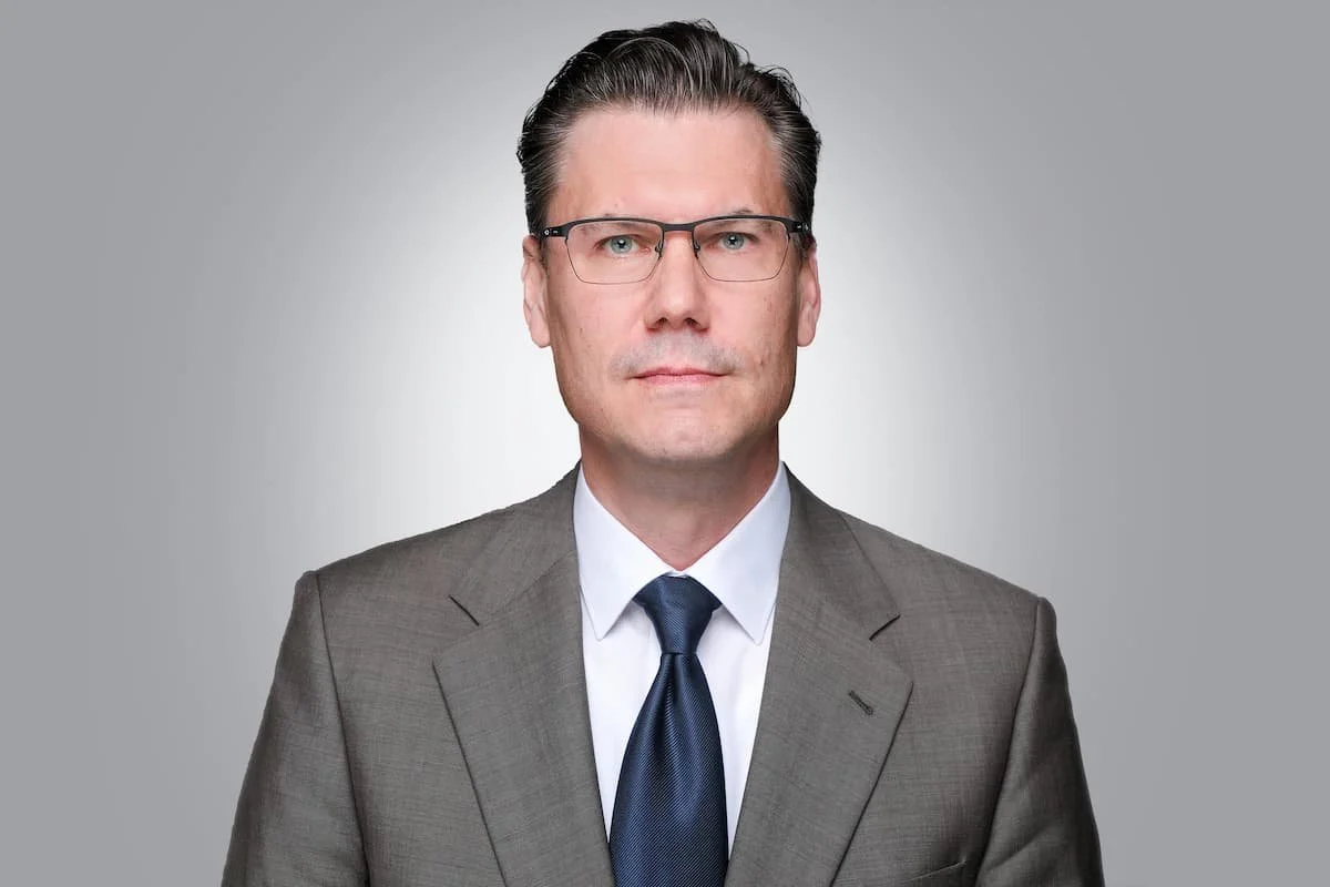 Porträt eines Mannes in einem grauen Anzug, weißen Hemd und dunkler Krawatte, mit kurzen, dunklen Haaren und Brille, vor einem hellgrauen Hintergrund.