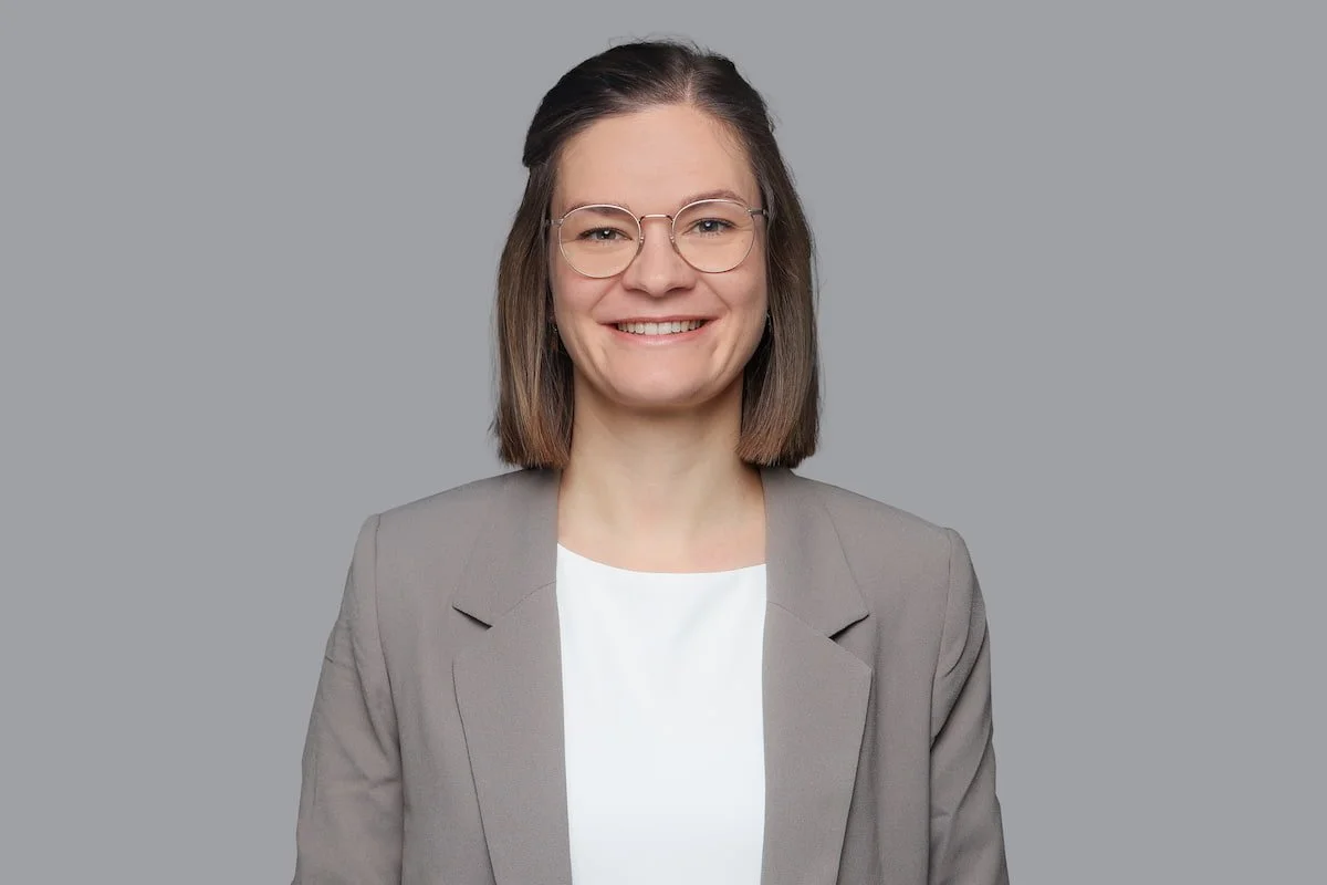 Professionelles Portrait einer jungen Frau mit Brille, braune Haare, grauem Blazer und weißem Hemd vor grauem Hintergrund.