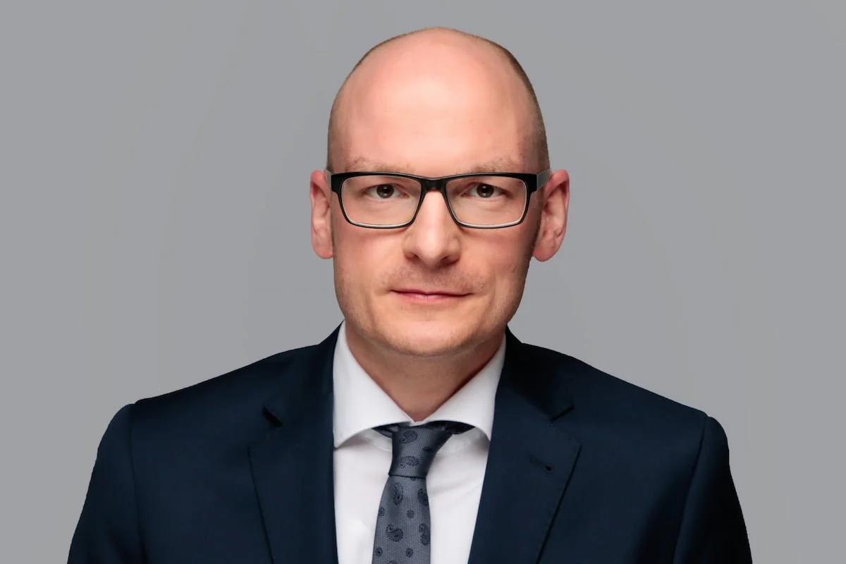 Porträt eines Mannes in Anzug mit Krawatte und Brille vor grauem Hintergrund.