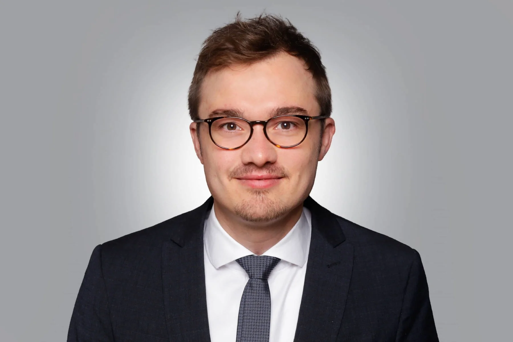 Porträt eines jungen Mannes mit Brille, dunklem Anzug, weißen Hemd und grauem Krawatte vor grauem Hintergrund.