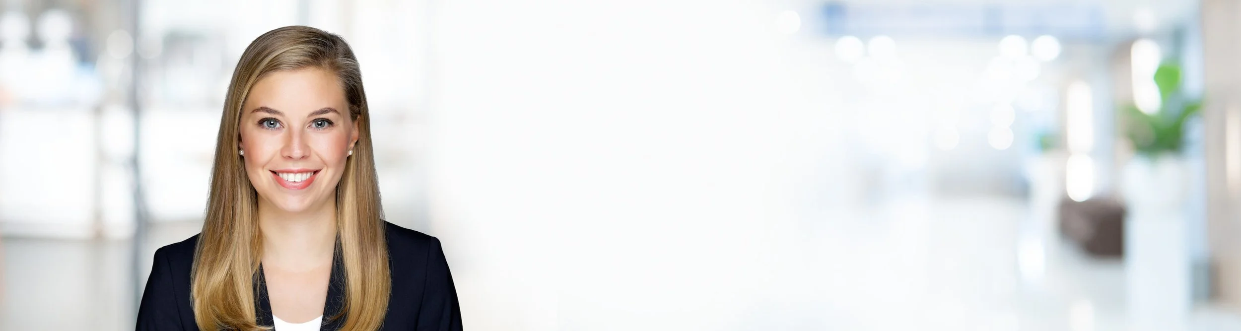 Lächelnde junge Frau mit blonden Haaren in einem schwarzen Blazer vor einem hellen, unscharfen Bürohintergrund.