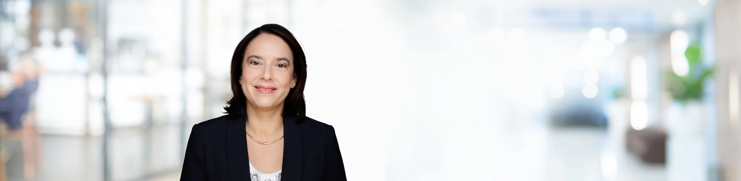 Porträt einer lächelnden Frau in einem schwarzen Blazer vor einem verschwommenen hell erleuchteten Hintergrund.