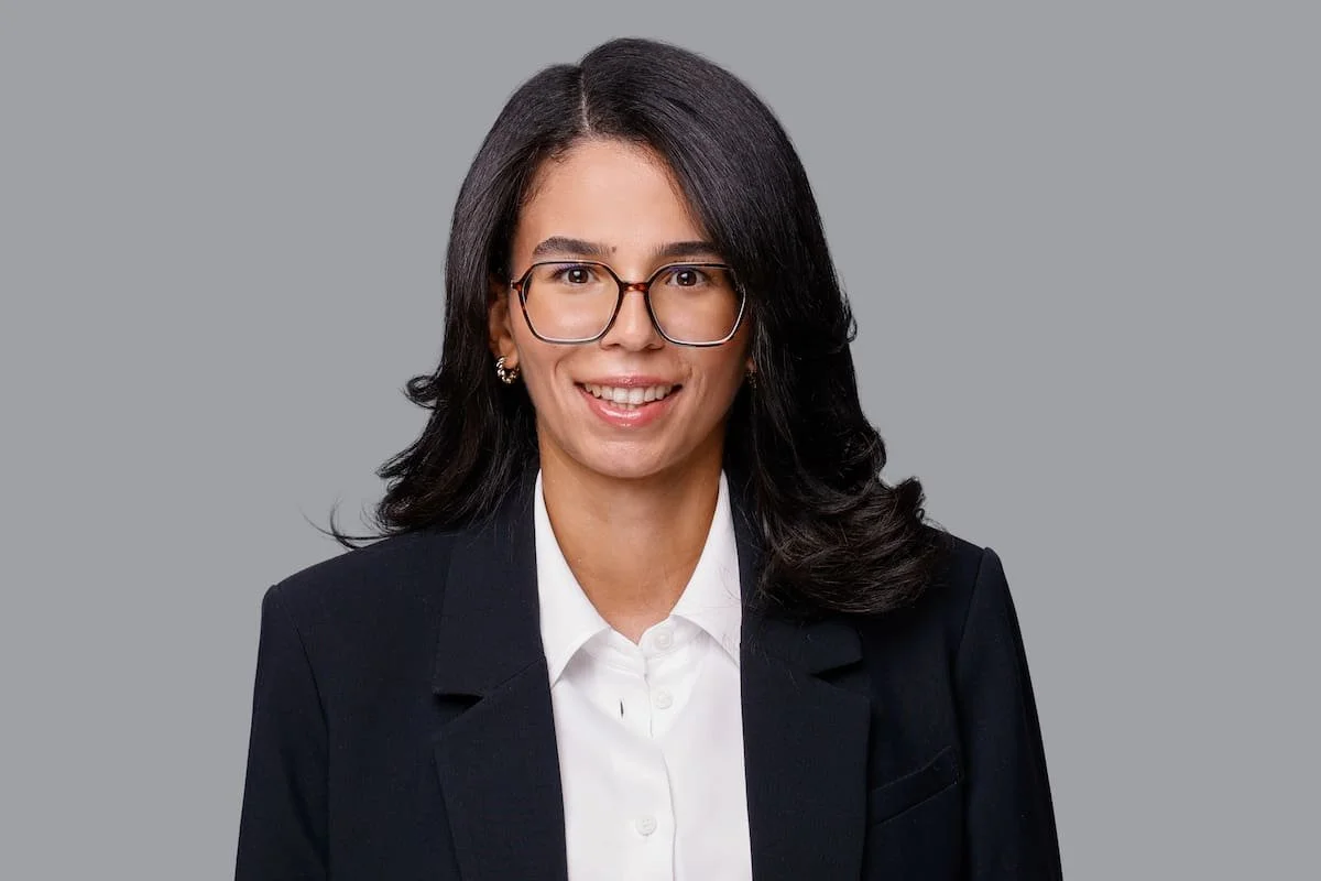 Porträt einer lachenden Frau mit schwarzen Haaren, Brille, in formeller Kleidung vor grauem Hintergrund.