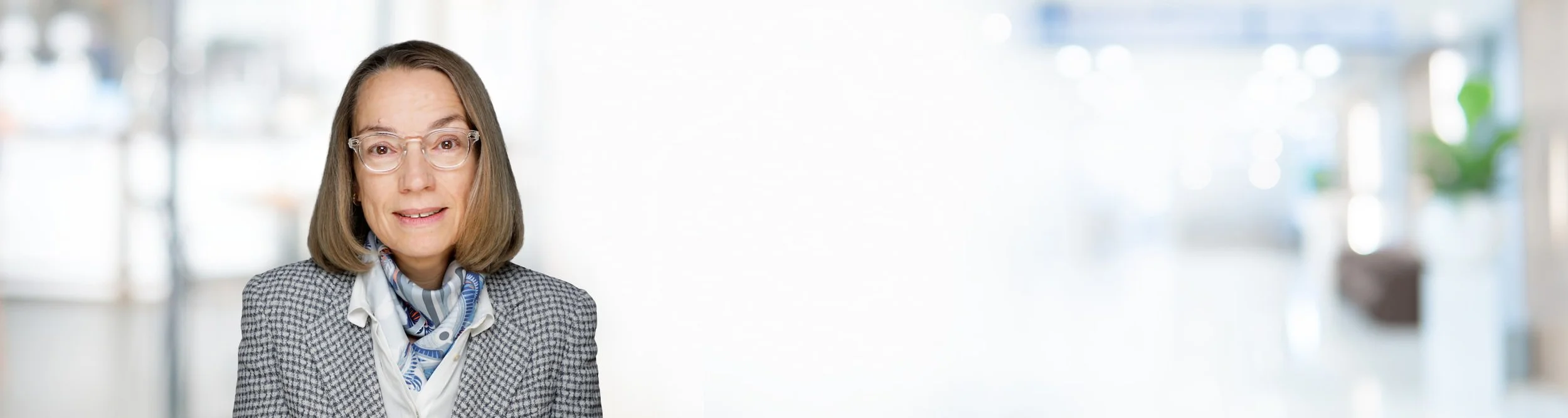 Eine mittelalterliche Frau mit kurze braune Haare, Brille, grauem Blazer und einem weißen Schal mit blauen Mustern. Sie steht in einem hellen, modernen Raum, der unscharf im Hintergrund ist.