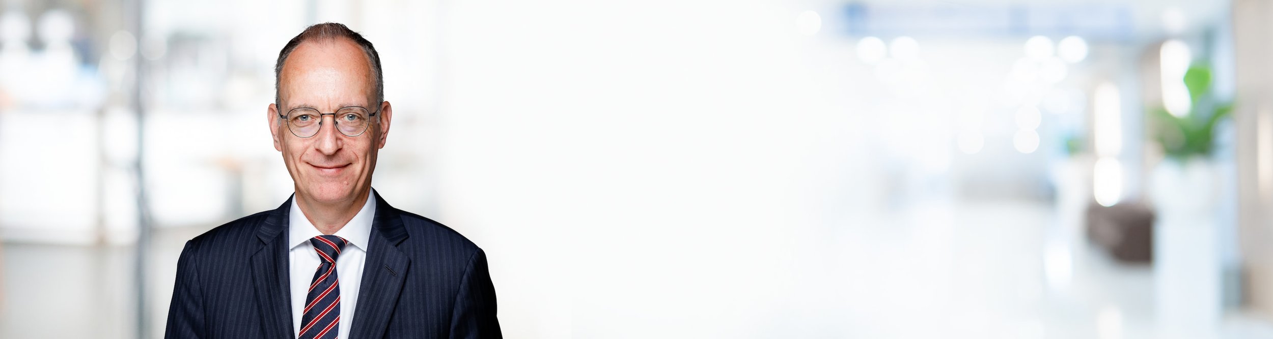 Ein älterer Mann in Anzug und Krawatte vor einem hellen, unscharfen Hintergrund in einem modernen Gebäude.