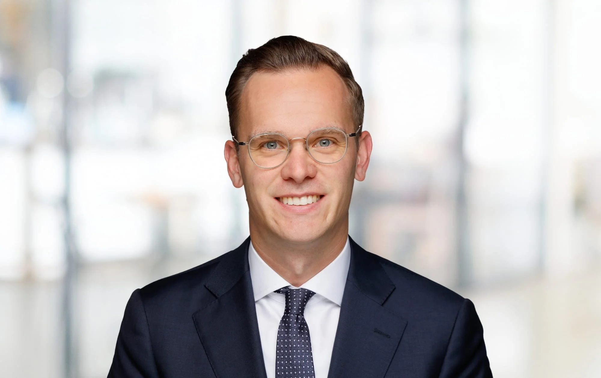 Porträt eines Mannes in Anzug mit Brille vor unscharfem Bürohintergrund.