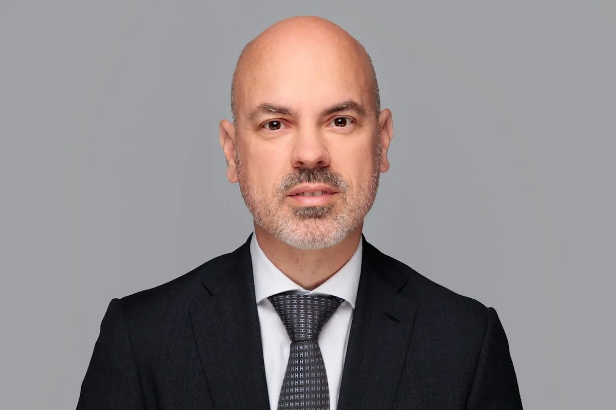 Porträt eines Mannes in formeller Kleidung mit Blond- und Grauhaar, er trägt einen schwarzen Anzug, weißes Hemd und eine schwarze Krawatte, gegen grauen Hintergrund.