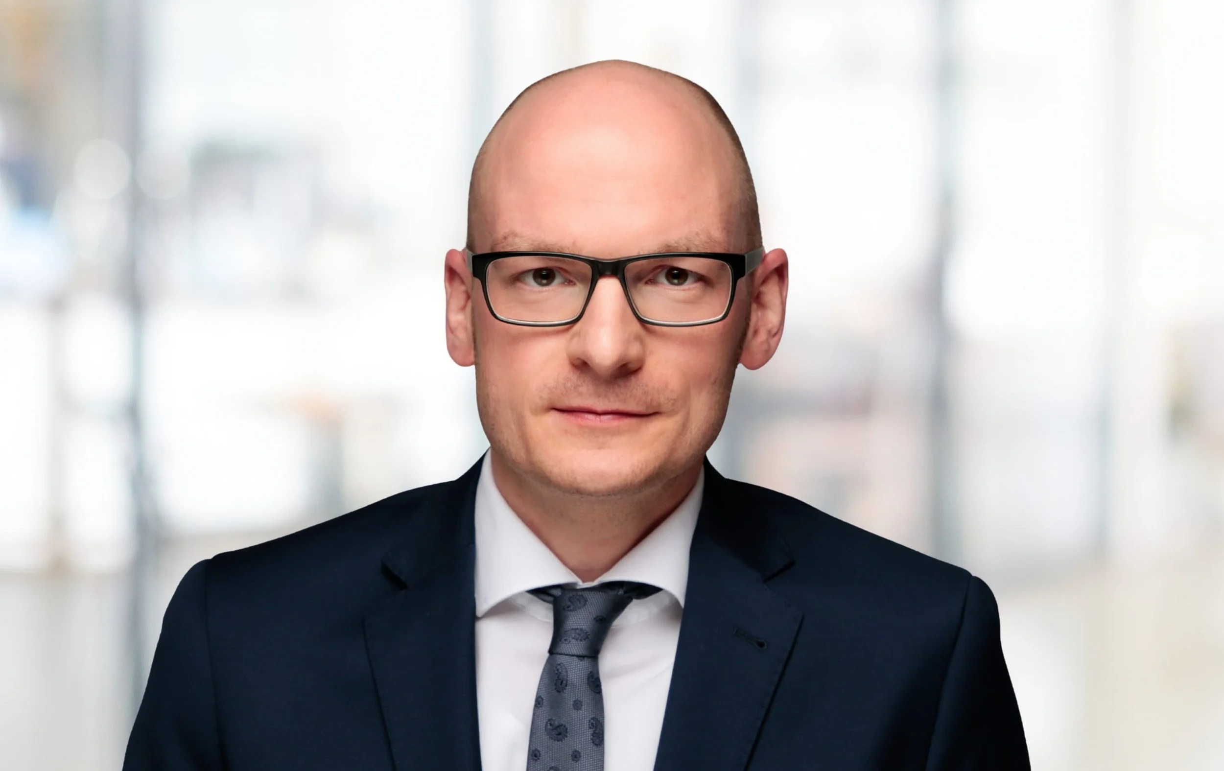 Porträt eines Mannes in Anzug, Krawatte und Brille vor unscharfem hellen Hintergrund.